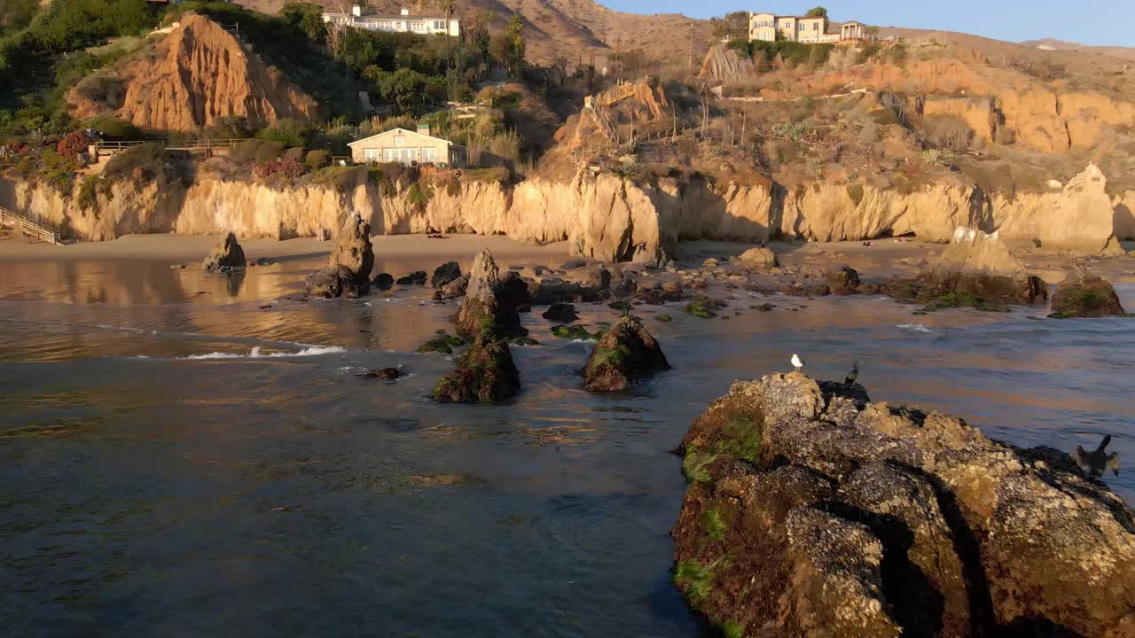 vista aérea de las rocas a la orilla de la playa el matador en malibu california, pájaros parados sobre las rocas y olas rodando en la orilla durante la hora dorada, hermoso paisaje