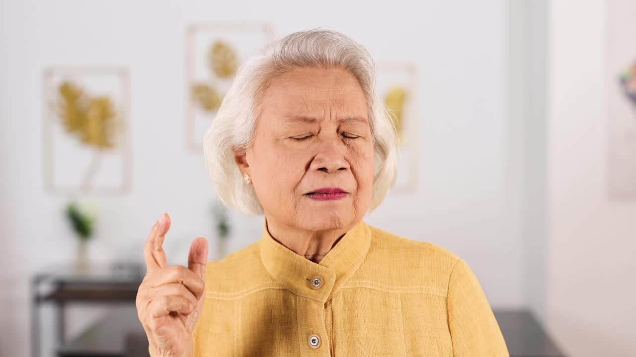 Senior Asian woman in yellow top thinking thoughtfully in bright, modern living room, soft lighting