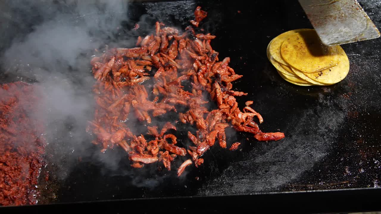 Vista dall'alto di carne al pastor che sfrigola su una griglia calda al rallentatore. Il vapore sale in modo spettacolare in questo ambiente street food all'aperto, catturando le texture, l'olio sfrigolante e il saporito processo di cottura