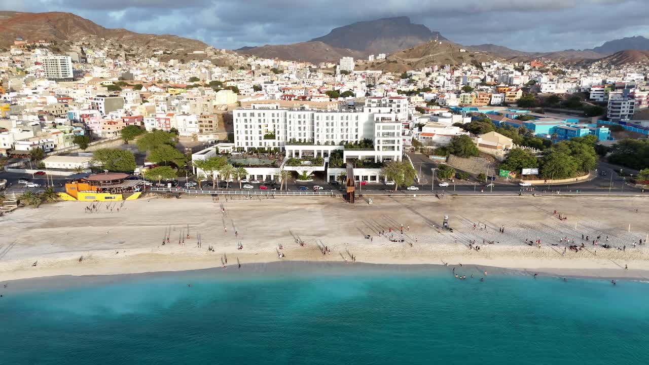 Widok z lotu ptaka na plażę Laginha w Mindelo, Sao Vicente, Republika Zielonego Przylądka. Turkusowe fale oceanu uderzające o piaszczystą plażę z miastem i wulkanicznymi górami w tle.