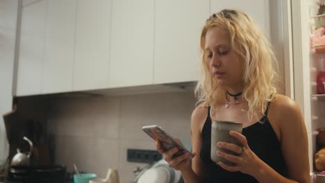 a tired blonde girl in a black top drinks tea in the morning and looks at her phone in the morning in the kitchen near the refrigerator