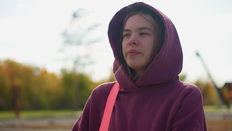 fitness enthusiast in burgundy hoodie walking on urban street adjusting pink side bag strap on shoulders one hand in trouser pocket among modern buildings and autumn foliage under bright daylight