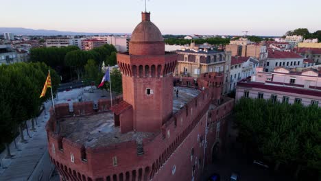 clip de avión no tripulado rodeando la fortaleza de le castillet en perpignan, francia por encima al atardecer, con un músico tocando en el techo y la bandera francesa volando en la suave brisa