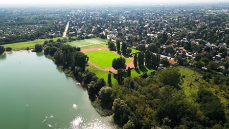 Erkunden-Sie-Mit-Der-Drohne-Eine-Sonnenverwöhnte-Sommerlandschaft:-Ein-Weitläufiges-Feld,-Einen-Ruhigen-See,-Badegäste,-Einen-Belebten-Basketballplatz-Und-Ein-Lebhaftes-Fußballfeld