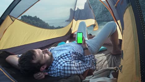 the solar panel attached to the tent. the man sitting next to mobile phone charges from the sun. a young guy lies in a camp tent, holding a smartphone with a green screen. camp tent stands on beach.