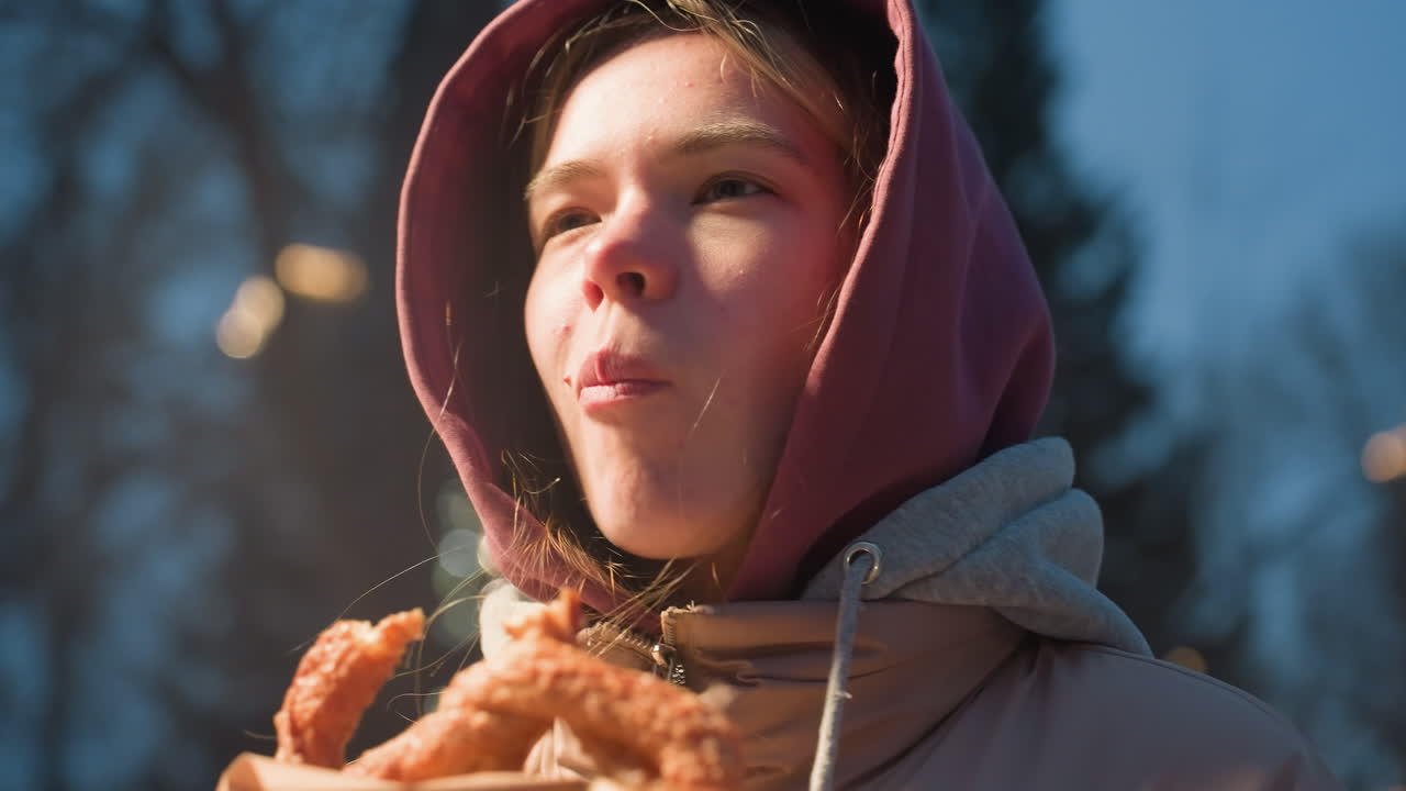 primer plano de una mujer disfrutando de un bocadillo con la luz iluminada destacando su cara en un entorno urbano de invierno, parque al aire libre nevado en el fondo