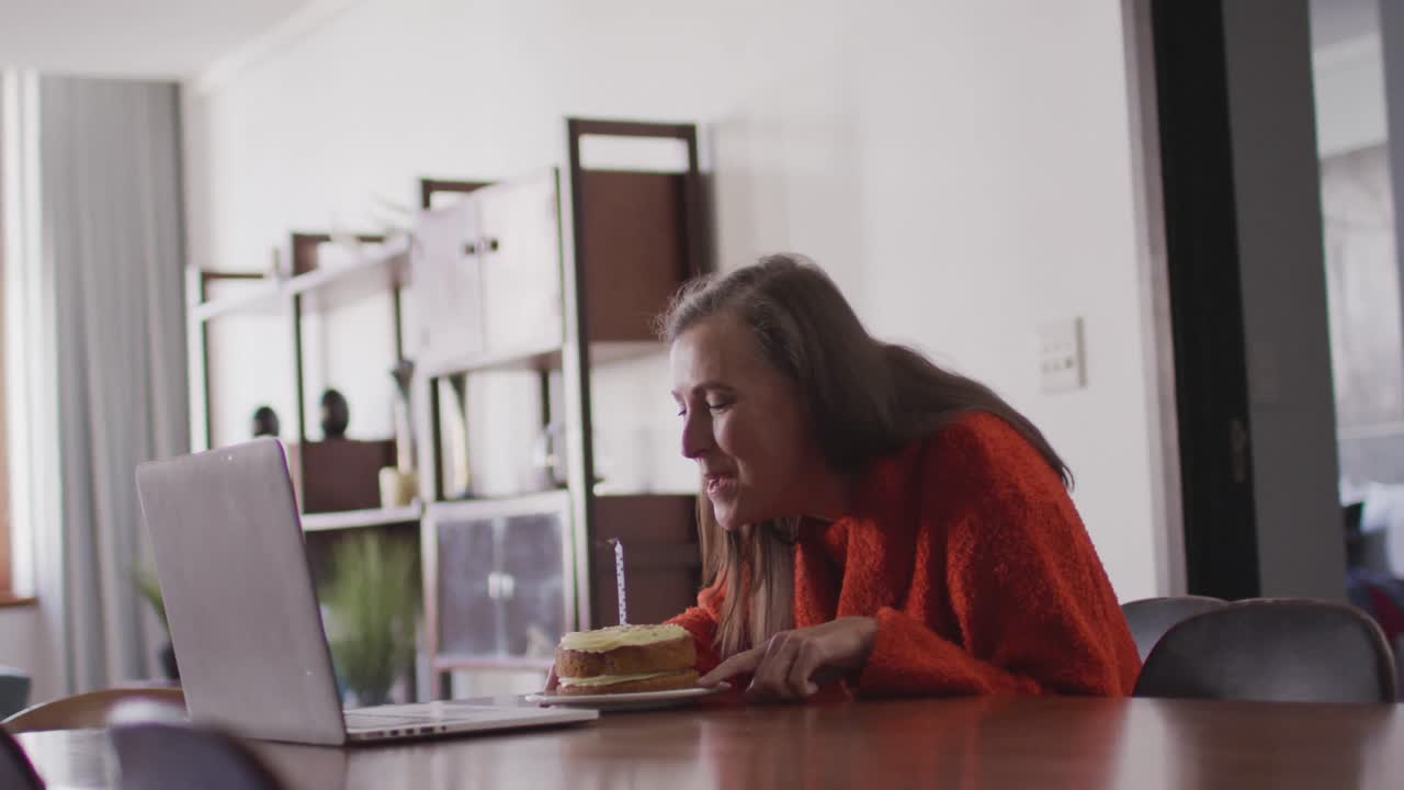 Woman blowing candle on the cake while having a video chat on her laptop at home