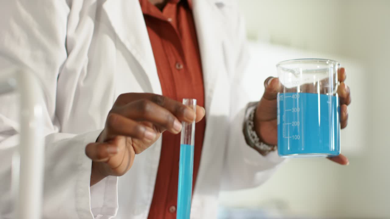 Upon lifting test tube, scientist is steadying beaker and tube while comparing blue liquid at bench