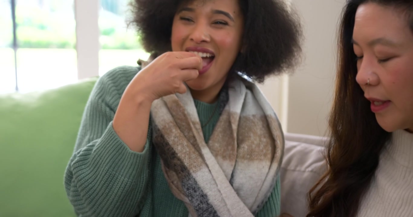 Mulheres afro-americanas e asiáticas no sofá passando uma tigela branca de pipoca, pegando e comendo enquanto assistem TV