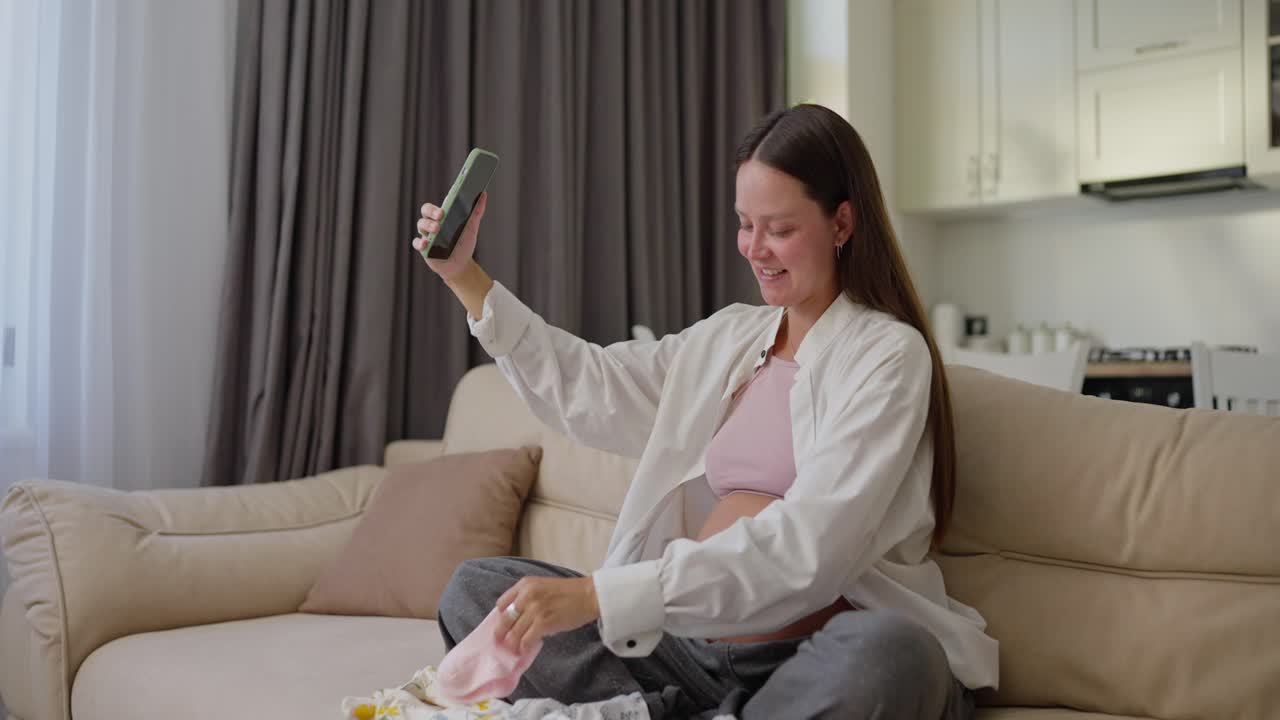 행복한 임신 한 여성은 임신에 대해 블로그를 작성하고 소파에 집에서 작은 아기 옷을 보여주는 아기를 기대합니다.