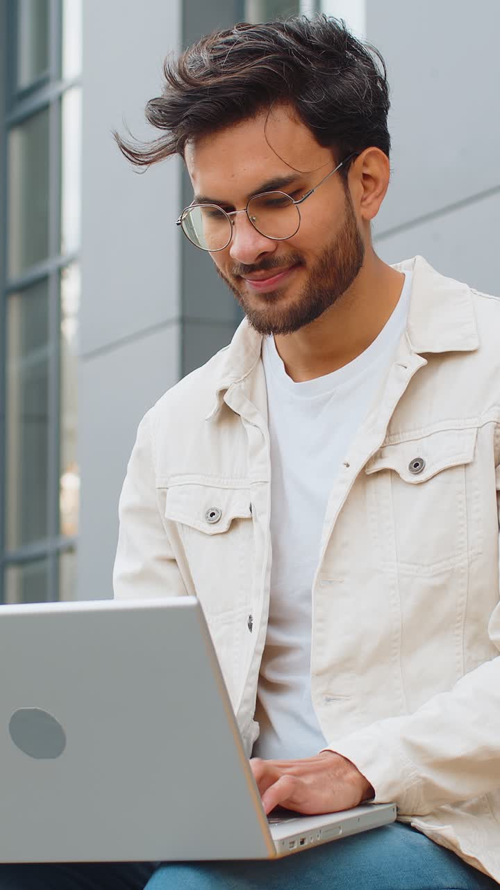 Indian man freelancer working on laptop computer sends messages reading email outdoors on bench