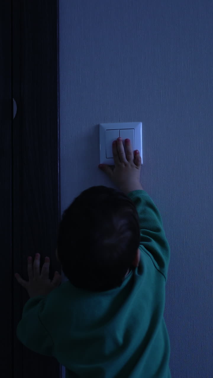Small baby boy comes up to the light switch in the dark room. Beautiful toddler turns on the light and walks away happily. Vertical video