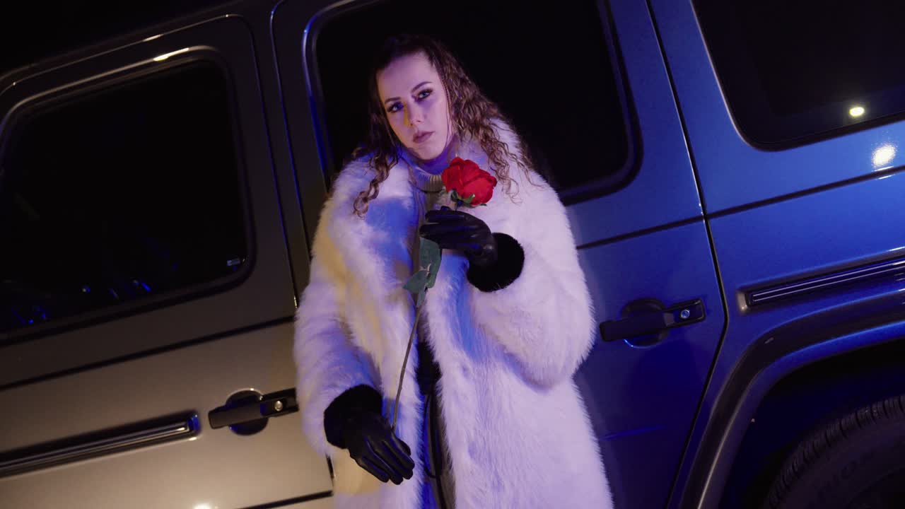 Nattbilde av en stilig kvinne i en hvit pels og skinnhansker som holder en rød rose foran en luksuriøs SUV. Konsept for romantikk, Valentine-innhold, luksuriøs livsstil
