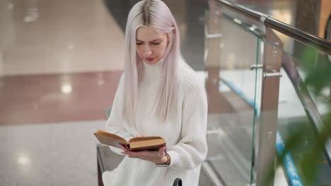 high angle view of woman seated on mall bench turning book pages with focused gaze wearing white sweater and jeans black bag beside modern railing escalator background soft lights retail environment