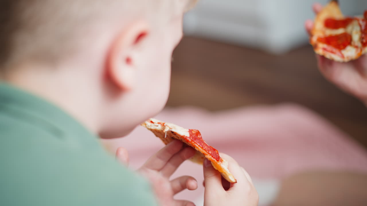 Rear view of little boy in green shirt eating slice of pepperoni pizza indoors while another hand holding pizza is seen in background, with soft cozy lighting