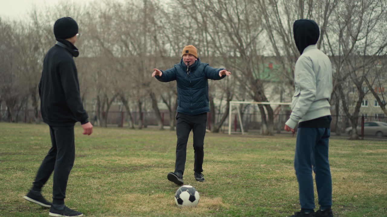 A coach sets the ball in the middle of two players facing each other as he blows the whistle, the background features a goal post, trees, and a car passing by