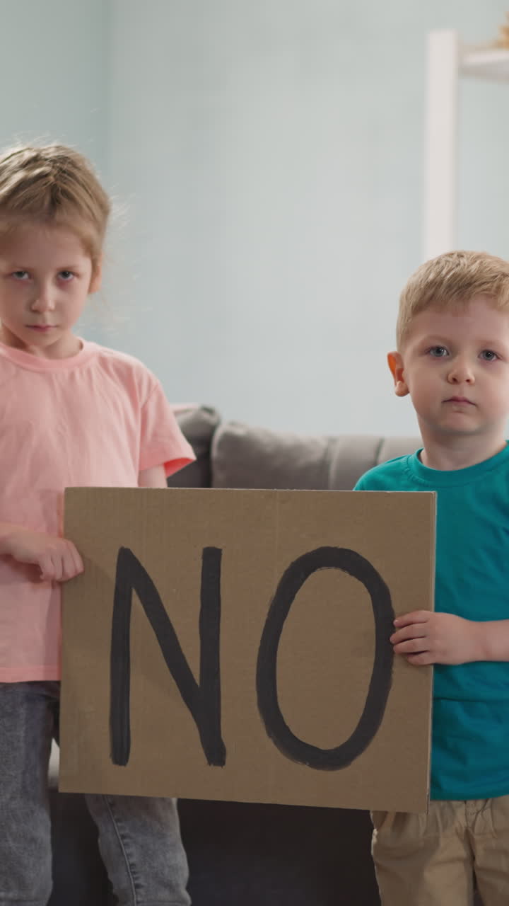 Unhappy little children show placard with word NO standing in living room slow motion. Kids protest against negative events in world. Message presenting