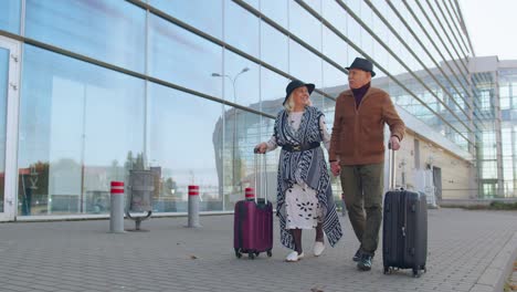 senior pensioner tourists grandmother grandfather walking from airport hall with luggage on wheels