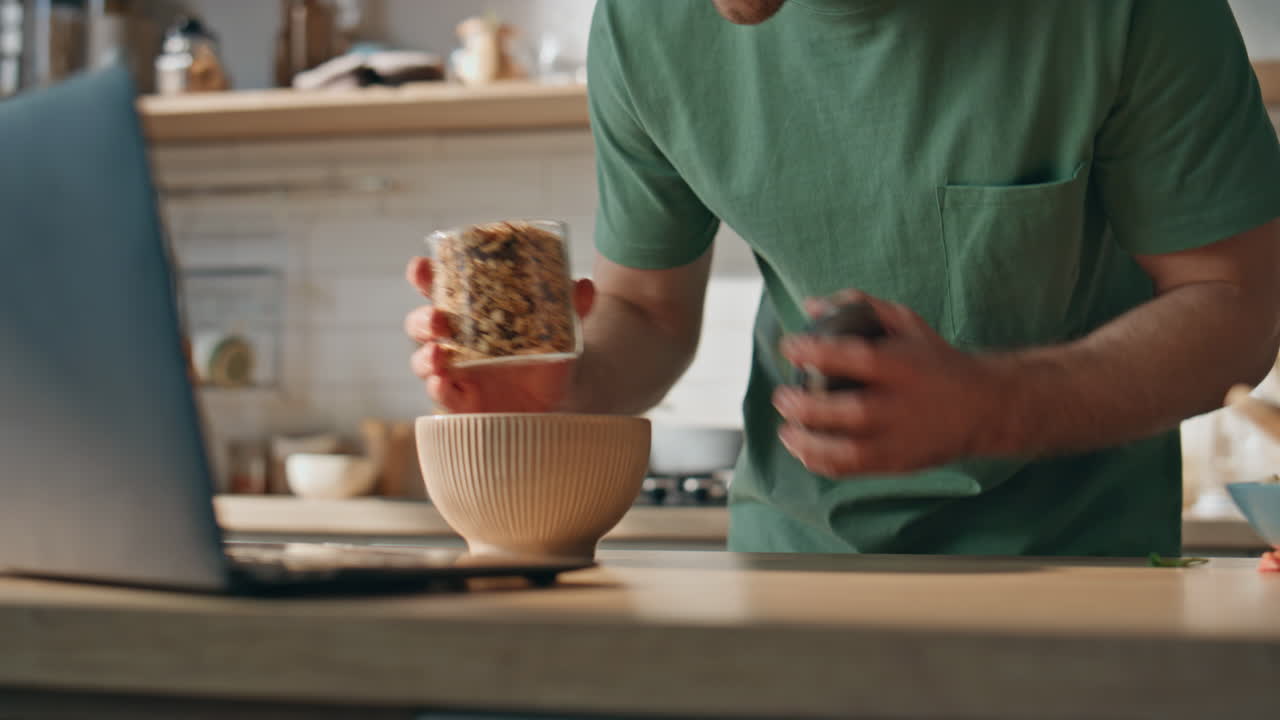 Serious brunette pouring granola in plate at flat closeup. Man watching laptop
