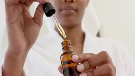 happy biracial woman wearing bathrobe and holding bottle of oil in bathroom, slow motion