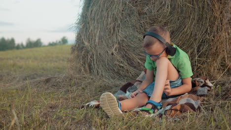 boy in green shirt and glasses sits on blanket outdoors, headphones on, focused on music, resting head on knee while scratching leg, against vast farmland backdrop