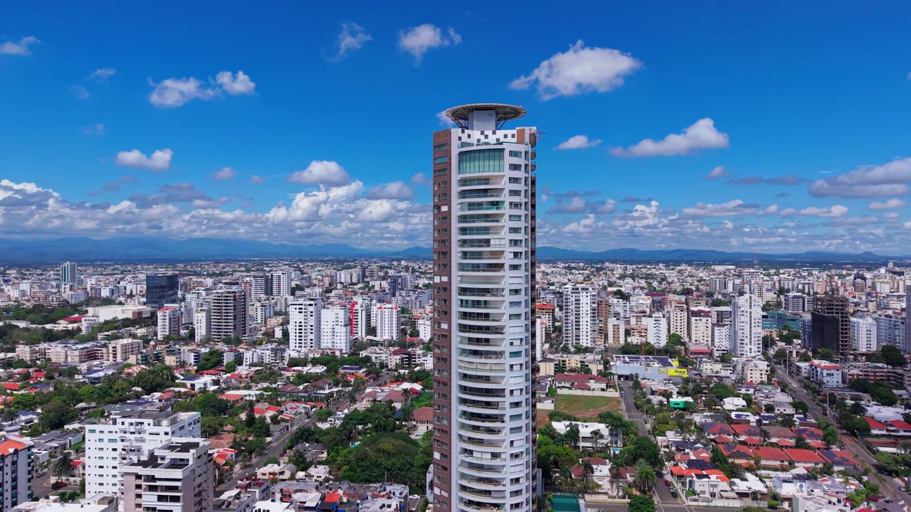Uma tomada aérea de drone subindo verticalmente sobre um parque verde denso, revelando um arranha-céu residencial moderno e alto e o horizonte da cidade em expansão.