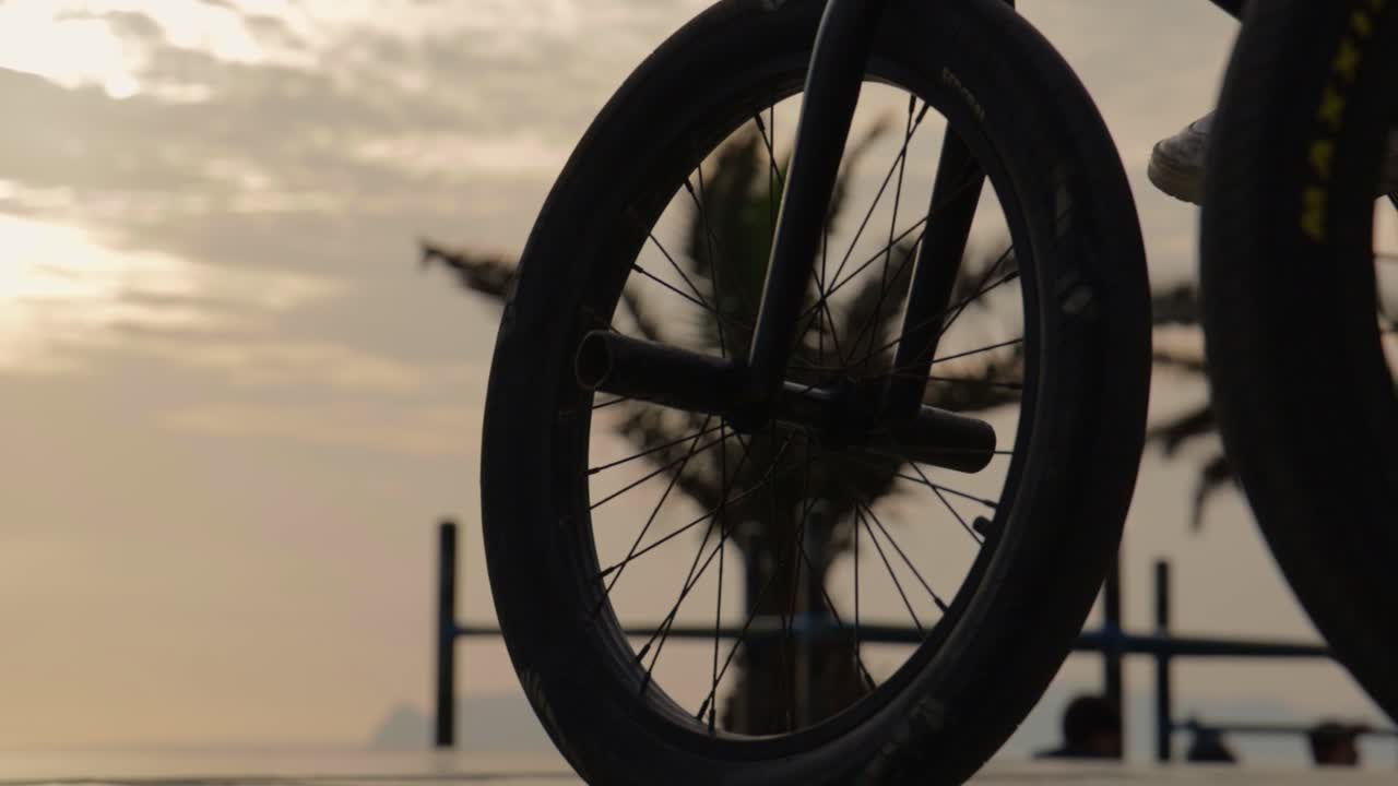Primo piano della ruota di una bici BMX con persone che vanno in bici e sullo skateboard al tramonto