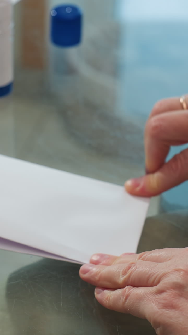Manos doblando papel sobre una mesa de cristal, primer plano de unas manos adultas arrugando y alineando hojas cerca de una barra de pegamento; movimientos cuidadosos y firmes creando un pequeño folleto para un proyecto de manualidades casero, luz natural brillante.