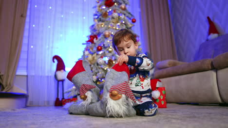 beautiful caucasian baby boy sits on the floor under the christmas tree. kid pulls a candy from the stocking and rejoices. low angle view.