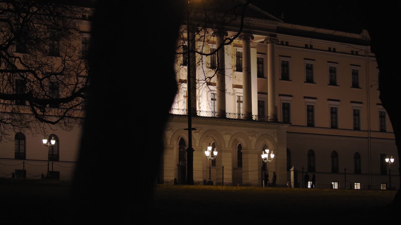 Stabilized wide to close-up 4K shot of dark trees moving in parallax motion in front of the illuminated Norwegian Royal Palace with armed guard walking in front of building, at night in Oslo Norway