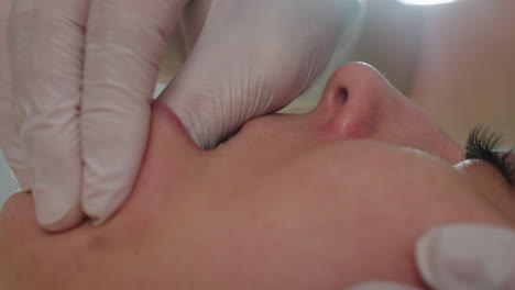 close up of visitor lying face up on massage chair with eyes closed and mouth open gently while wellness technician wearing gloves dips fingertip inside mouth and massages cheek under clinical light