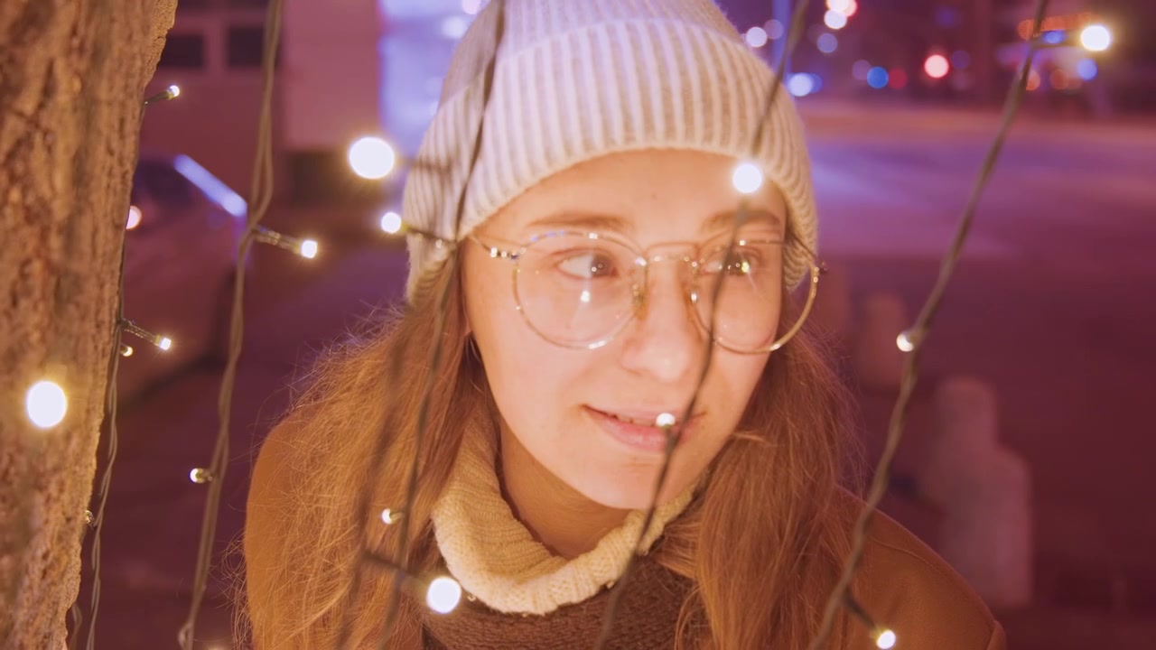 Junge kaukasische Frau unter Lichtern, trägt Mütze und Brille, lehnt an einem Baum auf einer winterlichen Stadtstraße, warmes Bokeh-Leuchten, nachdenkliches Lächeln, festliche Abendatmosphäre, sanfte Kinotöne, funkelnd.