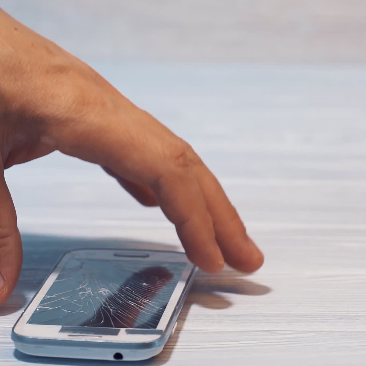 Slow motion of a man's hand taking broken smartphone from the floor. Cracked screen of an expensive cellphone is lying on the wooden floor inside.