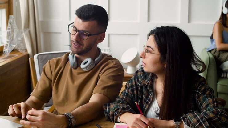 la telecamera si concentra su uno studente con le cuffie che parla con la compagna utilizzando il laptop, quindi mette a fuoco gli oggetti sul tavolo