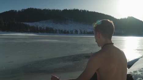 joven sin camisa sentado al lado de un lago de montaña congelado en invierno con nieve en el paisaje y luz solar brillante