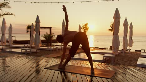 A-young-guy-does-yoga-and-takes-different-positions-of-his-body-at-the-dawn-of-the-sun-on-a-sunny-beach-in-the-morning.-Grows-on-a-sunny-beach-covered-with-planks