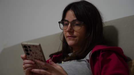 bored woman with glasses chatting on smartphone while sitting on sofa
