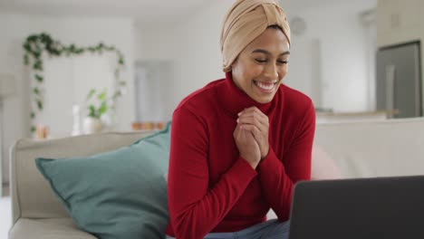 Video-of-happy-biracial-woman-in-hijab-sitting-on-sofa-and-having-video-call