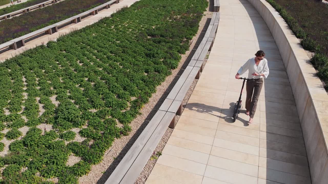 atractiva mujer caucásica está caminando con un scooter en una hermosa ciudad moderna en un día soleado, vista aérea. transporte ecológico moderno, popular entre los jóvenes