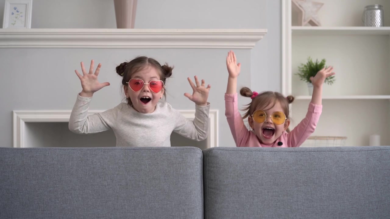 Two Excited Little Girls Wearing Sunglasses Popping Up Behind a Sofa