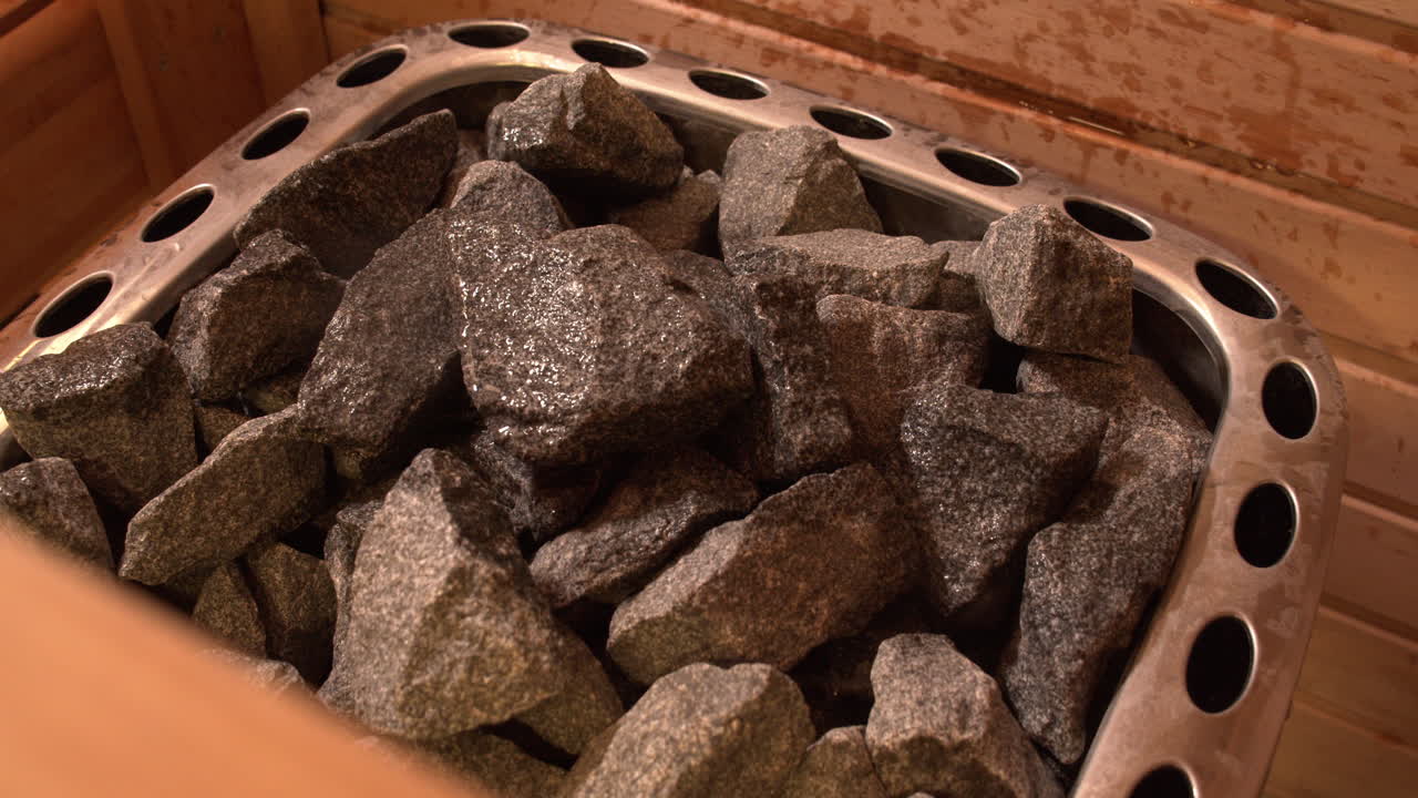 나무 사우나 내부의 뜨거운 돌에 물이 튀는 모습을 보여주는 클로즈업 샷으로 증기를 생성합니다.