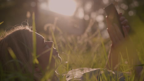 close-up of young woman lying on grassy field, using her phone while bathed in warm sunlight, the natural light creates a glowing effect around her