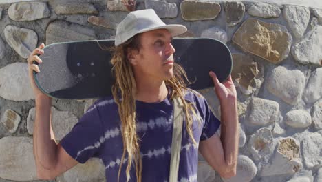 video of smiling caucasian man with dreadlocks holding skateboard in the sun by stone wall