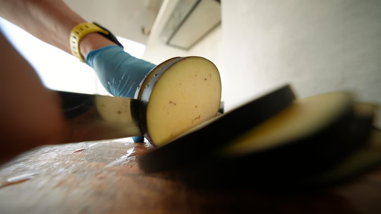 Hånd i blå handske skiver aubergine på træskærebræt