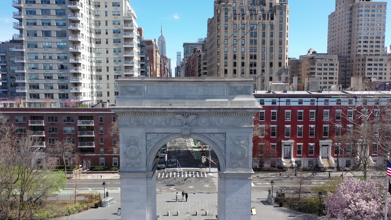 An aerial view of the Washington Square Arch in NYC. The drone camera pedestal down - dolly in towards the arch. The park - the surrounding streets are mostly empty - it's a bright - sunny day.