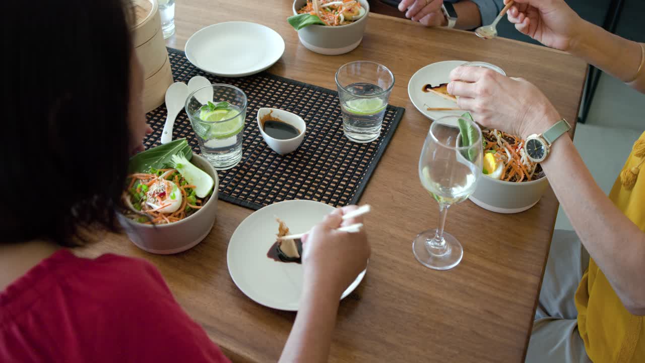Diverse female friends reaching for chopsticks, sharing noodle bowls, dipping sauces at wood table