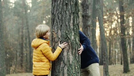 Fröhlicher-Kaukasischer-Vater-Und-Sohn-Spielen-Im-Wald,-Verstecken-Sich-Hinter-Einem-Baumstamm-Und-Sehen-Sich-Von-Verschiedenen-Seiten-An