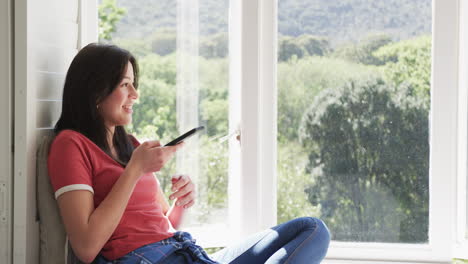 Mujer-Birracial-Feliz-Sentada-Junto-A-La-Ventana-En-Una-Habitación-Soleada-Hablando-Por-Teléfono-Inteligente,-Espacio-Para-Copiar,-Cámara-Lenta