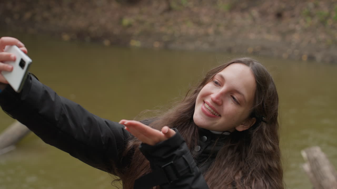 White woman taking selfie by river during overcast autumn walk black jacket, long brown hair, gentle smile, blowing kiss, playful pose for camera, calm water and muted trees in background conveying