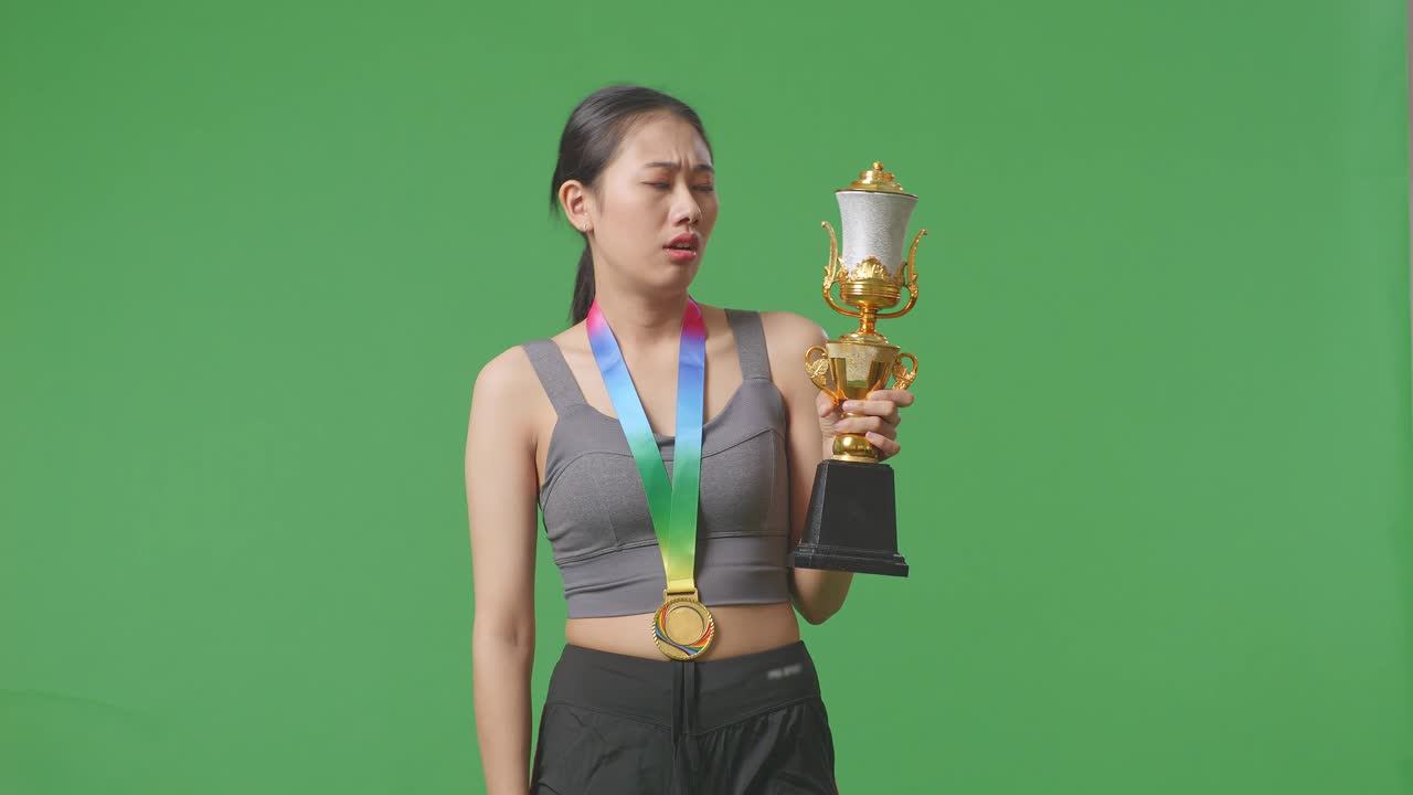 금메달을  아시아 여성은 트로피를 보고 머리를 흔들며 불만을 품고 스튜디오의 녹색 스크린 배경에서 첫 번째 우승자로 승리합니다.